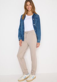 Světle béžové tepláky se bočními pruhy, kombinované s bílým tričkem a modrou džínovou bundou. Klasické pletené boty s modrými akcenty.