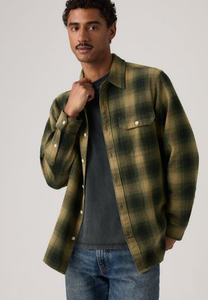 Homme aux cheveux bouclés et à la moustache portant une chemise à carreaux verte et noire déboutonnée par-dessus un t-shirt sombre et un jean bleu, debout devant un fond uni.
