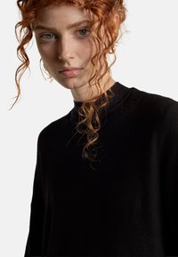 Schwarzer Pullover mit geripptem Ausschnitt, der eine weiche Textur und eine entspannte Passform aufweist. Das Modell hat lockiges rotes Haar, das das Gesicht umrahmt, vor einem neutralen Hintergrund.