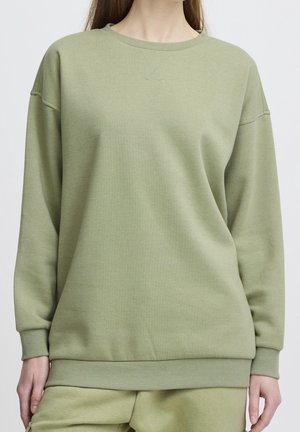 Jeune personne portant un sweatshirt vert clair oversize et un pantalon assorti, debout devant un fond clair et uni.