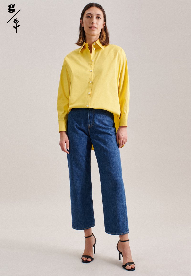 Frau in einem lockeren gelben Button-Up-Hemd, blauen weitem Jeans und schwarzen hochhackigen Sandalen, stehend vor einem hellen Hintergrund.