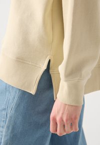 Maglione in cotone beige con polsini a coste e orlo laterale spaccato, abbinato a denim azzurro chiaro, con attenzione alla texture e alla vestibilità.