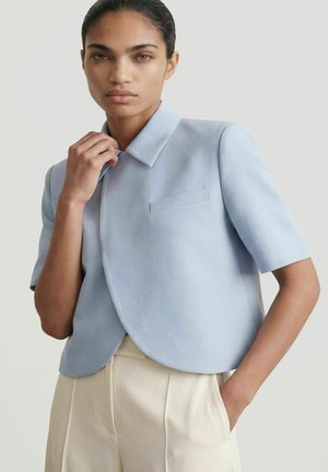 Žena s tmavými vlasmi v bledo modrej krátkej bunde a krémových nohaviciach s vysokým pásom stojaca s jednou rukou v vrecku a druhou pri golieri.