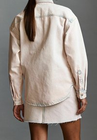 Chemise en jean oversize rose clair avec un ourlet incurvé, des poignets boutonnés et des bords effilochés sur une mini-jupe assortie. Texture douce, style décontracté.