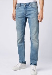 Hombre con jeans ajustados de color azul claro y zapatillas blancas, de pie contra un fondo liso claro.