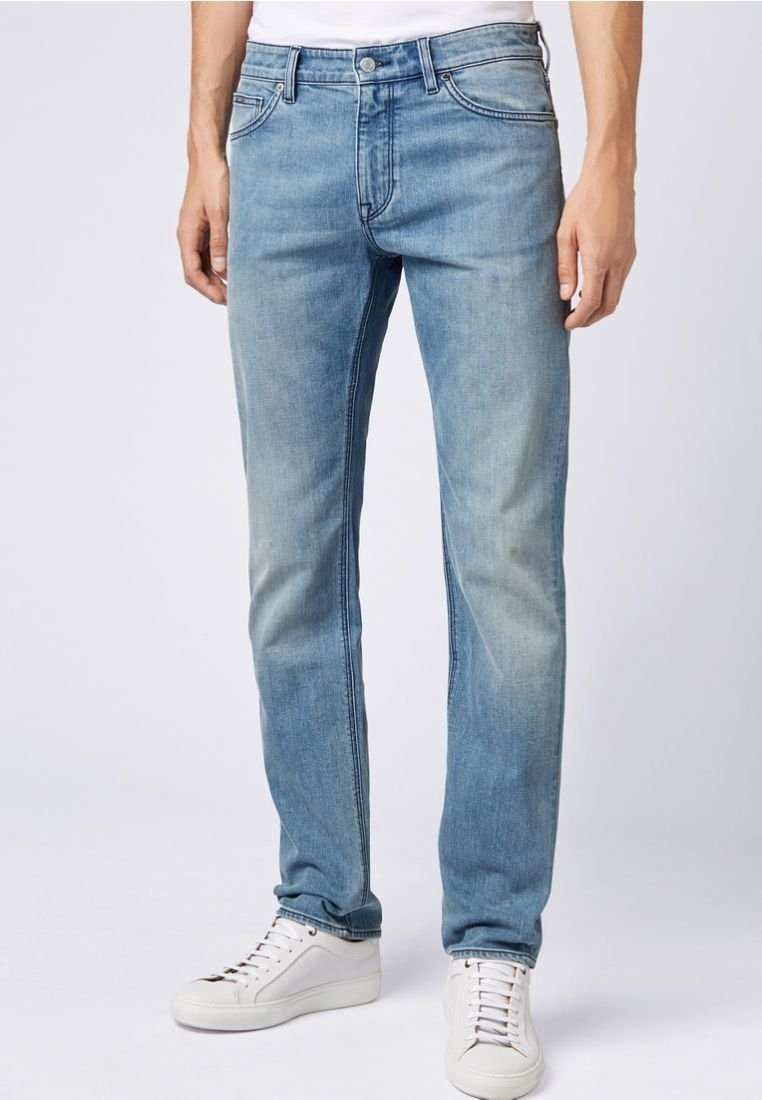 Hombre con jeans ajustados de color azul claro y zapatillas blancas, de pie contra un fondo liso claro.