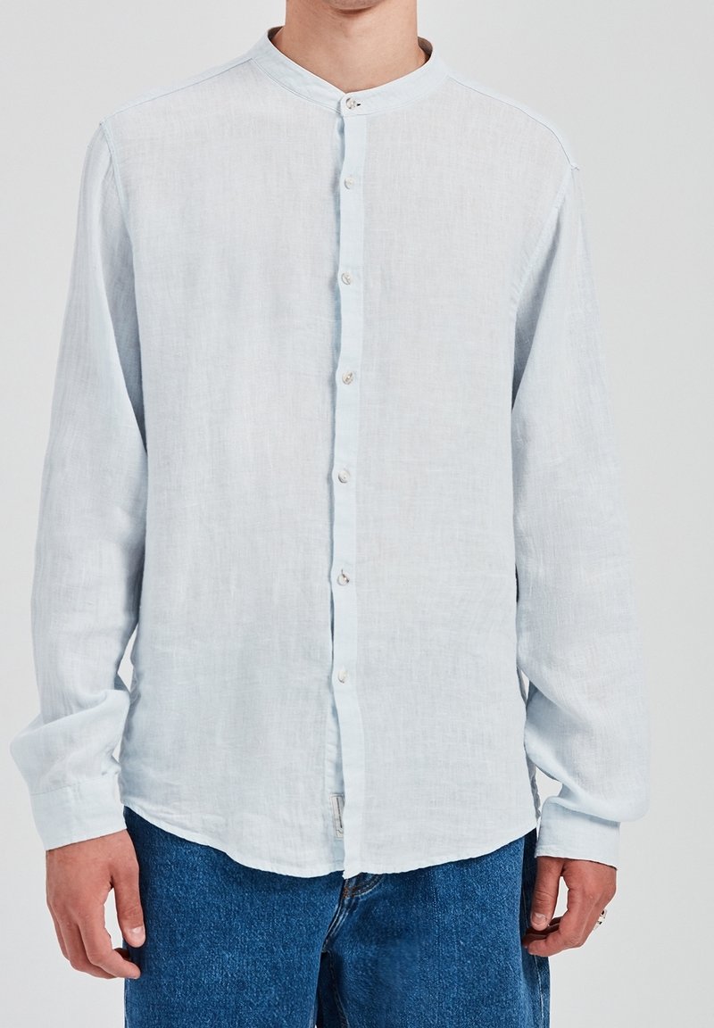 Jasnoniebieska lniana koszula z długim rękawem, z kołnierzykiem mandaryńskim, zapięciem na guziki i luźnym krojem. Gładka faktura i lekko przezroczysty materiał.