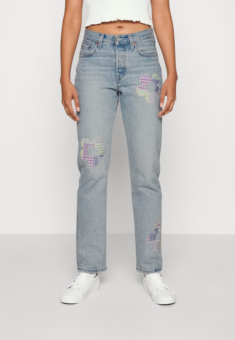 Persona que lleva vaqueros de mezclilla azul claro con parches de flores de cuadros coloridos en las piernas y zapatillas blancas, de pie contra un fondo simple.