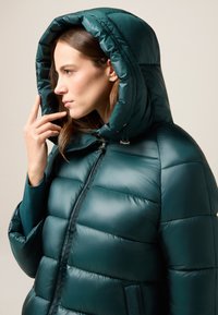 Teal pufferjack met een grote, gevoerde capuchon, voorzien van een ritssluiting, geborduurde textuur en zijzakken voor extra functionaliteit.