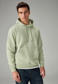 Sudadera verde clara de tela suave, con un bolsillo frontal, cordones ajustables y puños acanalados, combinada con jeans oscuros.