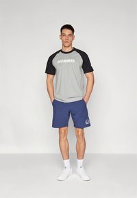 Hall halli t-särk mustade raglaanvarrukatega ja suure valge "HUMMEL" tekstiga, siniste spordipükstega, millel on logo, ning valgete tossudega.