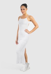 Donna che indossa un vestito maxi attillato grigio chiaro senza maniche con spacco laterale, abbinato a scarpe da ginnastica bianche, in piedi davanti a uno sfondo semplice.