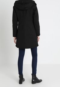 Czarna wodoodporna parka z kapturem, długimi rękawami i prostym krojem. Posiada szew na plecach i jest zestawiona z dopasowanymi niebieskimi dżinsami oraz czarnymi botkami za kostkę.