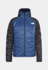 Blå och svart isolerad jacka med hög krage, framdragkedja och quiltad textur. Har varumärkeslogotypen på övre vänstra bröstet.