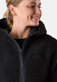 Schwarze Fleecejacke mit einer strukturierten, flauschigen Außenseite, Kapuze und Frontreißverschluss. Sie verfügt über ein Logo auf der Brust und robuste Reißverschlussbeschläge.