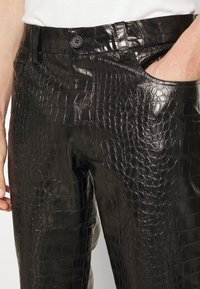 Nahaufnahme von schwarzen Hose aus Kunstkrokodilleder mit Knopf und einer Hand in der rechten Tasche, getragen mit einem weißen Oberteil.