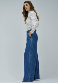 Mujer con blusa de encaje blanco de manga larga y pantalones vaqueros de tiro alto y pierna ancha de color azul con un cinturón de cadena, de pie de perfil con las manos en los bolsillos.