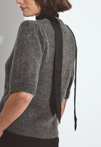 Pelēks adīts krekls ar īsām piedurknēm, kas aizmugurē ir sasiets ar melnu lentes. Mīksta tekstūra, brīva piegriezums un ribota apmale.