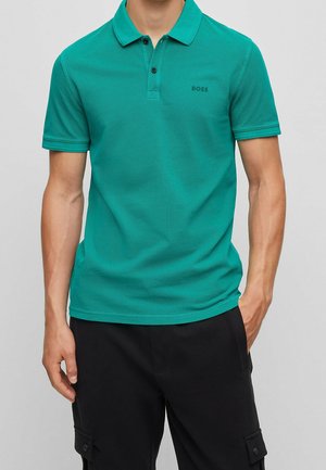 Man wearing teal short-sleeve polo shirt with black BOSS logo on chest and black cargo pants, standing against neutral background.