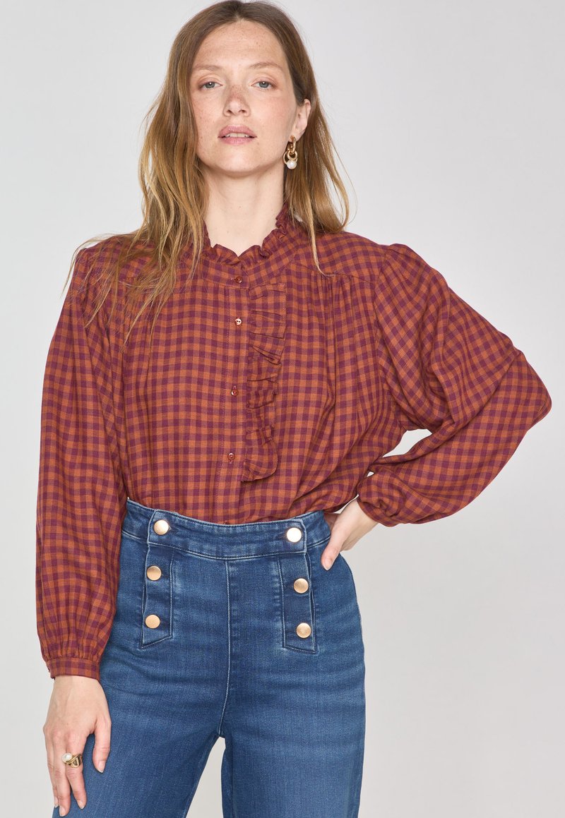 Blusa checkered marrone e bordeaux con colletto arricciato, caratterizzata da dettagli sui bottoni. Abbinata a jeans in denim blu a vita alta con bottoni in metallo.