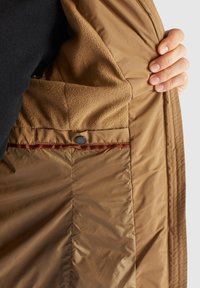 Main tenant une veste beige ouverte montrant la doublure intérieure en polaire et une poche intérieure avec fermeture par bouton sur fond neutre.