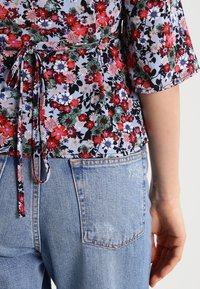 Blusa floreale in tessuto azzurro chiaro con fiori rossi, rosa e verdi. Presenta un lacciolo in vita e maniche a trequarti. Indossata con jeans in denim.