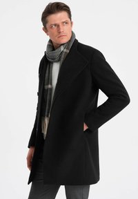 Abrigo de lana negro con grandes solapas, detalles de botones y bolsillos laterales, acompañado de una bufanda a rayas en gris y negro.