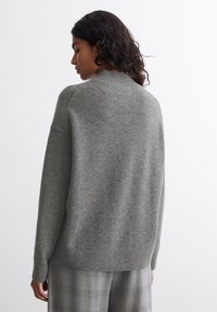 Pulover gri din tricot cu o croială lejeră, cu guler rotund și manșete cu nervuri. Textură moale cu un finisaj neted, purtat peste o pereche de pantaloni cu carouri.