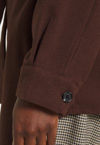 Hnědý vlněný kabát s texturovaným povrchem, s knoflíkem na manžetě v černé barvě. Ruka je částečně vidět, pod ní je kostkovaná látka.