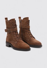 Botas de tobillo de ante marrón con un diseño de cordones en la parte frontal, cremallera lateral, correa ajustable con hebilla y un tacón bajo tipo bloque.