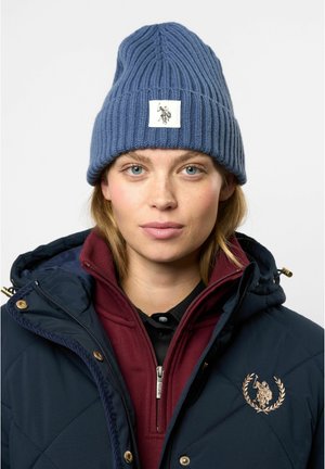 Blå ribbestrikket lue laget av ull, med en kvadratisk hvit lapp med en logo. Laget under en marineblå quiltet jakke og bordeaux genser.