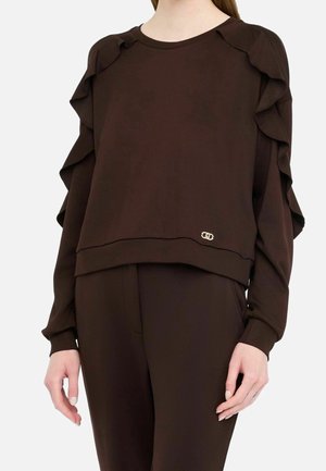 Personne portant un haut marron foncé à manches longues avec des détails à volants sur les manches et un pantalon marron foncé assorti sur un fond uni.