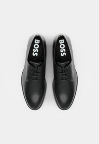 Chaussures habillées en cuir noir avec un bout arrondi, un design minimaliste, des lacets plats et une semelle intérieure foncée portant l'inscription "BOSS" en lettres blanches.