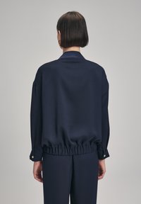 Blusa leggera col collare di colore blu navy con cintura elastica e polsini con bottoni. Tessuto liscio con design minimale, vista da dietro.