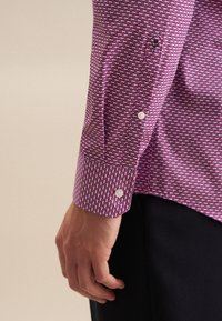Camicia con maniche lunghe caratterizzata da un motivo circolare rosa e viola, dotata di bottoni bianchi e un piccolo ricamo sul polsino.