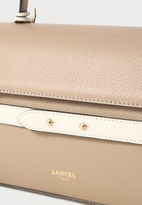 Tan leather handbag with a textured finish, featuring a white accent strap and gold hardware. Embossed Lancel Paris logo at the bottom.