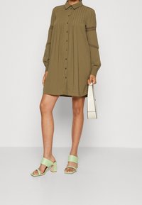 Khakifarbenes Hemdkleid mit Knopfleiste, langen Ärmeln und strukturiertem Stoff. Kombiniert mit mintgrünen Sandalen mit Blockabsatz und einer weißen Handtasche.