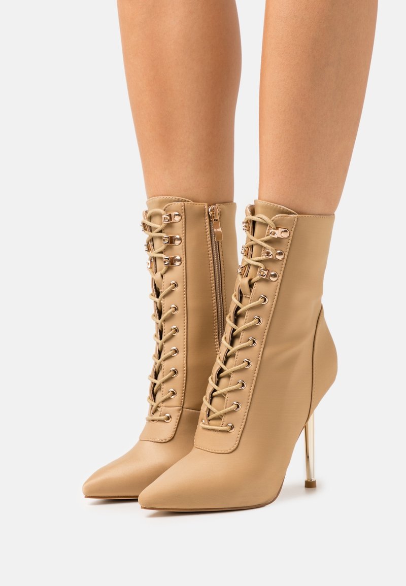 Bottes à talons hauts beige en matériau lisse, avec un bout pointu, un laçage à l'avant et une fermeture éclair dorée sur le côté.