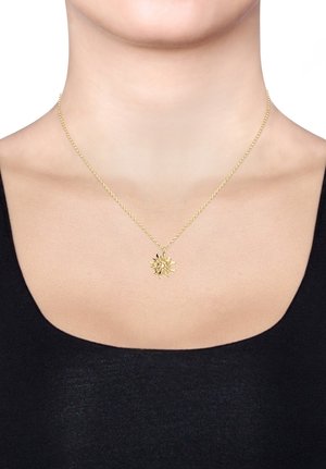 Goldene Halskette mit einem blumenförmigen Anhänger an einer gedrehten Kette, getragen auf nackter Haut mit einem schwarzen Oberteil.