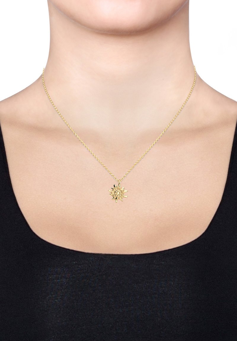 Goldene Halskette mit einem blumenförmigen Anhänger an einer gedrehten Kette, getragen auf nackter Haut mit einem schwarzen Oberteil.