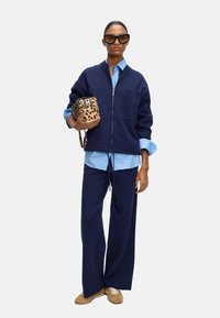 Navy-zipper-Sweater mit Taschen, kombiniert mit einem hellblauen Hemd und passenden weit geschnittenen Hosen. Hält eine Tasche mit Leopardenmuster und trägt dunkle Sonnenbrillen.