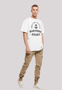 Weißes T-Shirt mit schwarzem Drucktext und Ankerdesign, kombiniert mit beigen Cargo-Hosen mit seitlichen Taschen und elastischen Bündchen.