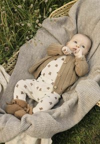 Cardigan beige lavorato a maglia sopra una tutina crema decorata con motivi di orsi marroni, abbinato a morbidi scarpini marroni, posato su coperte grigie dalle texture. 