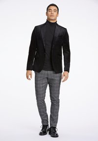 Zwarte fluwelen blazer met satijnen accenten, gecombineerd met een aangesloten zwarte coltrui en grijs geruite broek. Zwarte veterschoenen maken de look compleet.