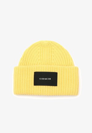 Geel gebreide beanie met een ribbeltextuur en een zwarte rechthoekige patch met witte tekst waarop "COACH" staat.