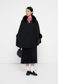 Cappotto di lana nera con colletto di pelliccia, fissato da una chiusura in argento. Abbinato a un top a maglia rosa, gonna plissettata, mocassini neri e una borsetta piccola.