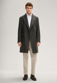 Donkergroene, dubbelgeknoopte wollen jas met grote knopen, zijzakken, gecombineerd met crèmekleurige geplooide broek en bruine enkellaarzen.