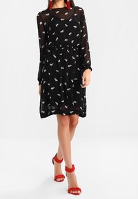 Vestido negro y transparente con mangas largas y cintura ceñida, con patrones de hojas blancas. Combinado con sandalias rojas de tiras esponjosas.
