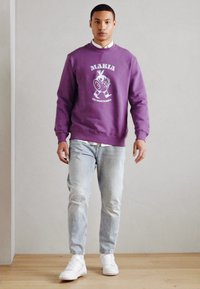 Lilla gensere med grafisk trykk, rund hals og ribbestrikkede mansjetter. Kombinert med lyseblå slitte jeans og hvite joggesko.