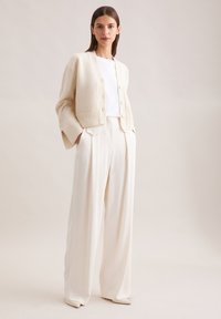 Gebreide, beige cardigan met een cropped ontwerp, voorzien van een knoopsluiting en ribgebreide manchetten. Gecombineerd met hoogsluitende, wijde witte broeken.